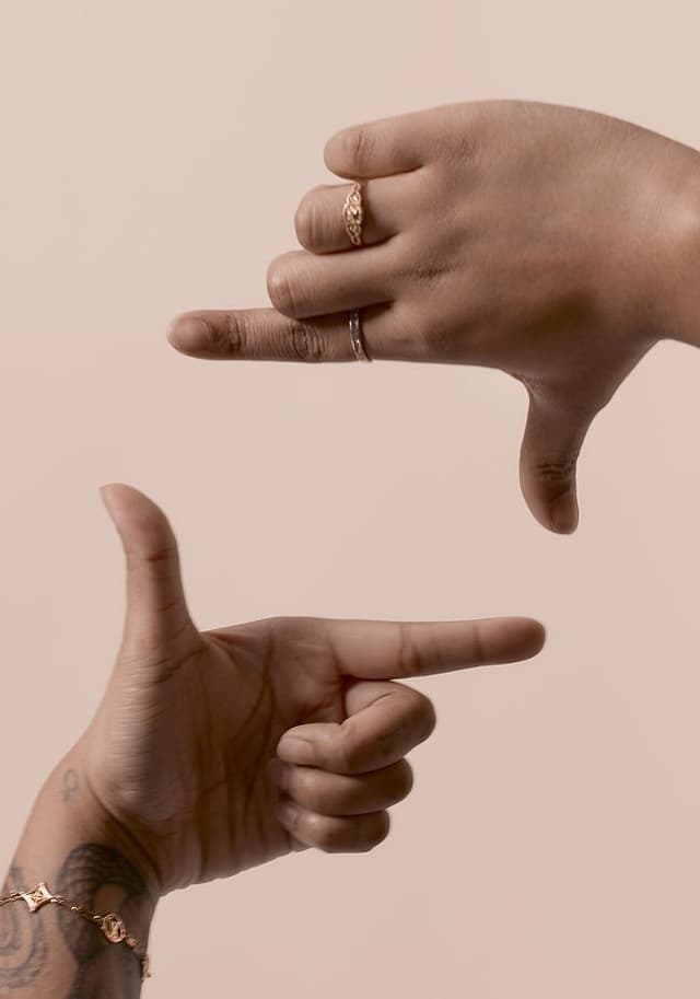 Close-up of two hands against a beige background making a rectangular frame shape with their thumbs and index fingers. One hand has tattoos and a bracelet, the other has two rings.