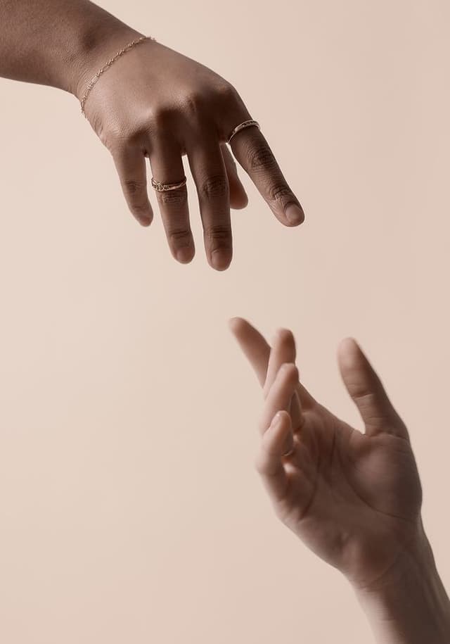 Two hands reaching towards each other against a neutral beige background, one hand adorned with rings and a bracelet, the other bare.