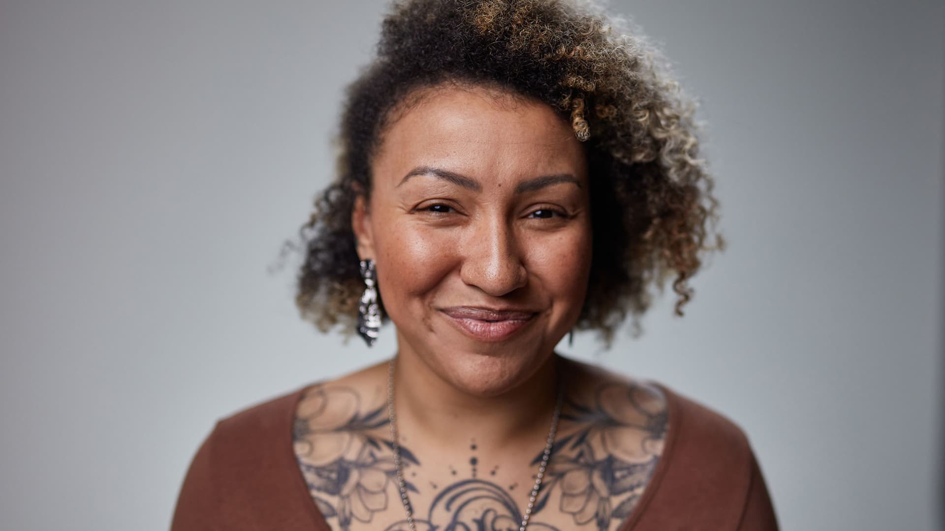 Close-up portrait of a smiling woman with curly hair, wearing a brown top, dangling earrings, and a visible floral chest tattoo.
