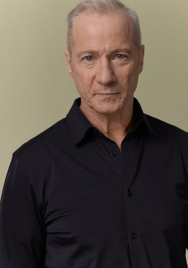 Portrait of an older man with short gray hair and blue eyes, wearing a black button-up shirt, against a plain light beige background.