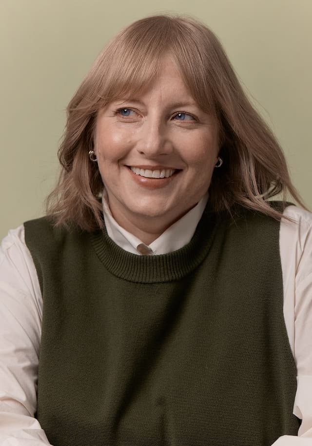 Portrait of a smiling woman with light brown hair wearing a white shirt and an olive green knit vest against a plain beige background.