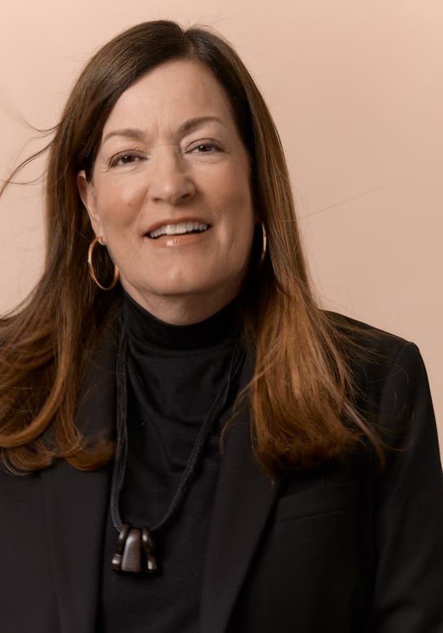 Portrait of a middle-aged woman with long brown hair wearing a black turtleneck, black blazer, hoop earrings, and a black pendant necklace, smiling against a beige background.