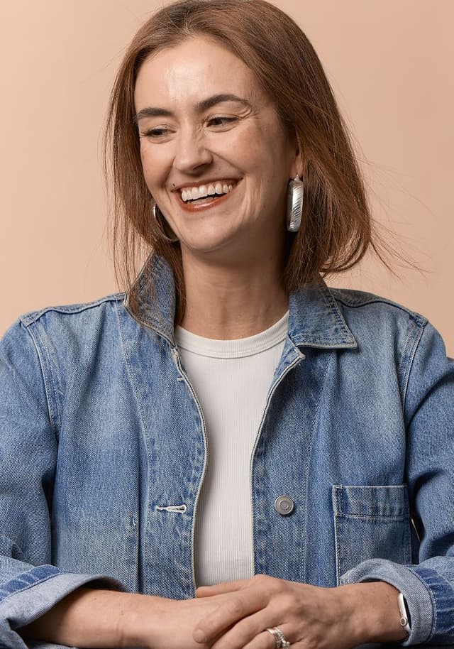 Smiling woman with brown hair wearing large silver earrings, a white shirt, and a blue denim jacket against a beige background.